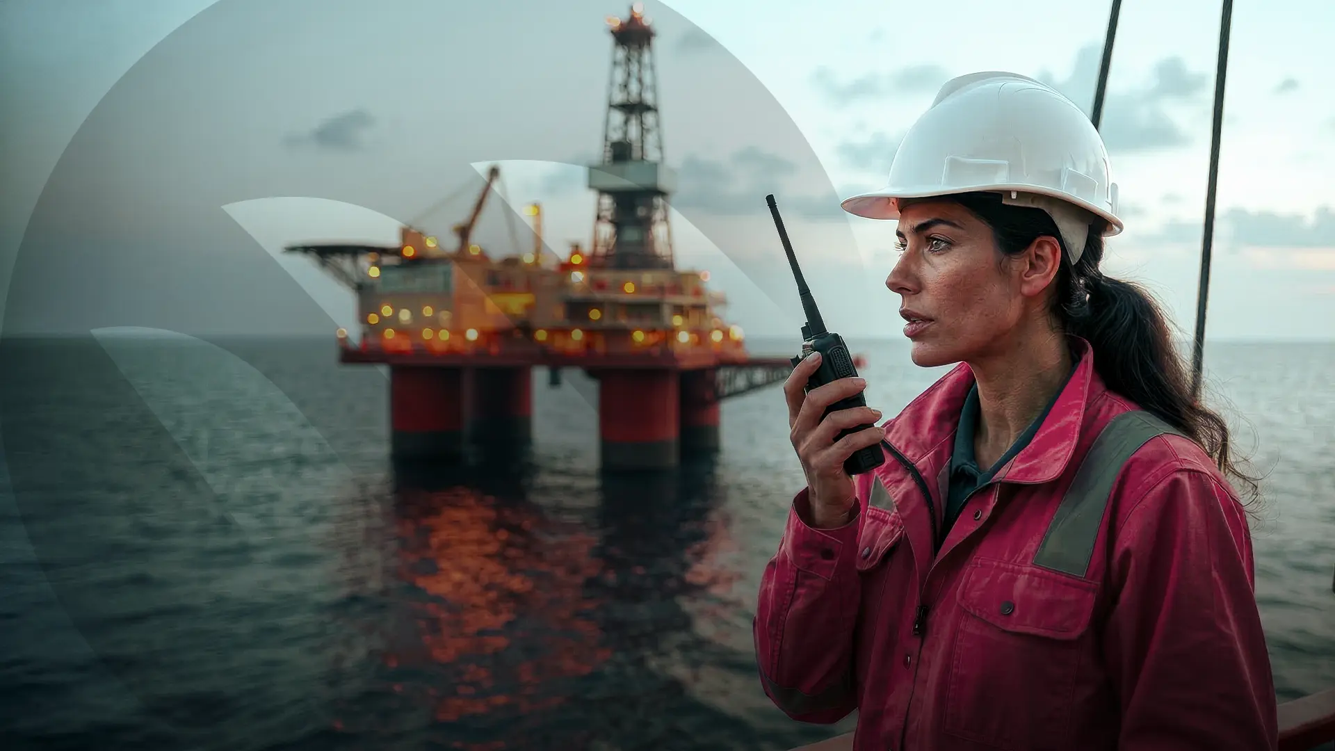 Mulher com uniforme vermelho e capacete branco fala em um aparelho de rádio, ao fundo uma plataforma de petróleo no oceano.